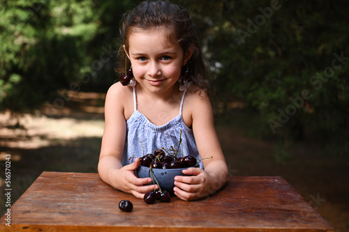 Child girl wearing ripe cherries earrings on her ears, standing by a table with a bowl of freshly picked cherry berr photo