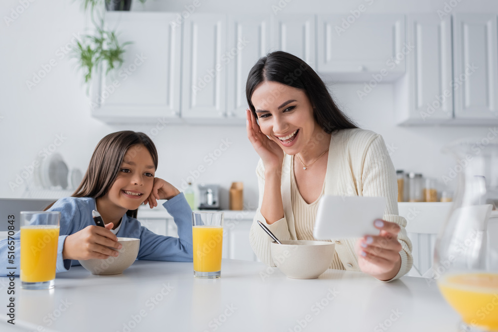 smiling nanny showing smartphone while having breakfast with girl in kitchen. smiling nanny showing smartphone while having breakfast with girl in kitchen.