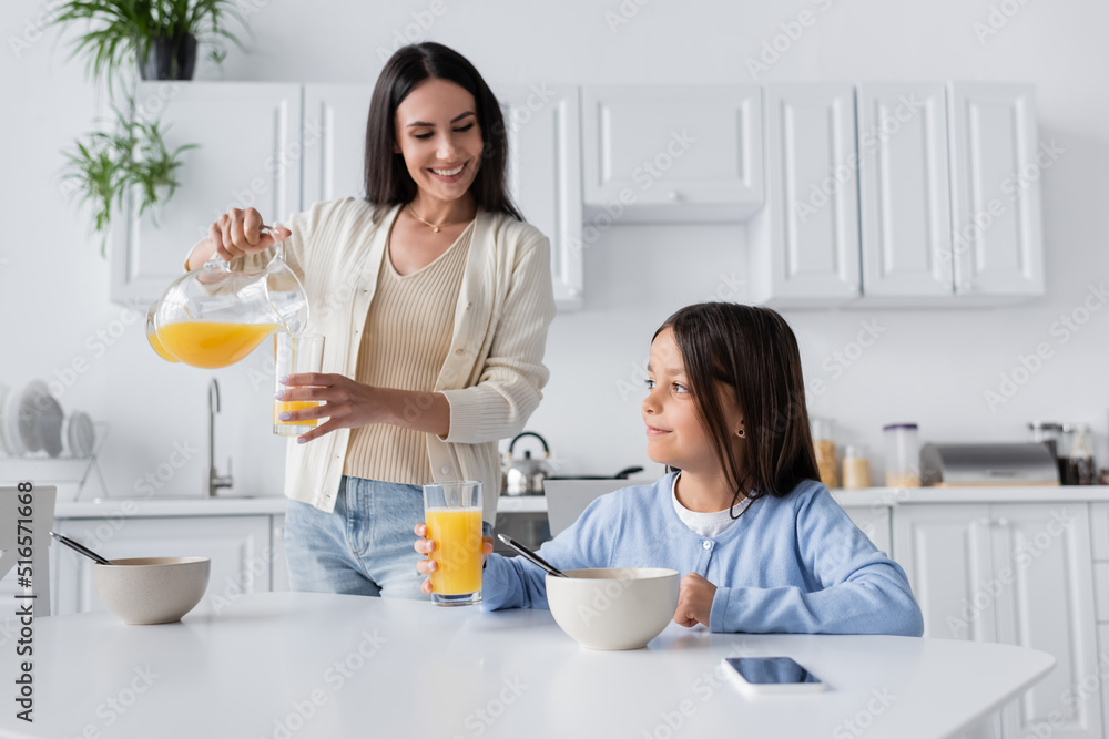 brunette nanny pouring orange juice near smiling girl having breakfast in kitchen. brunette nanny pouring orange juice near smiling girl having breakfast in kitchen.