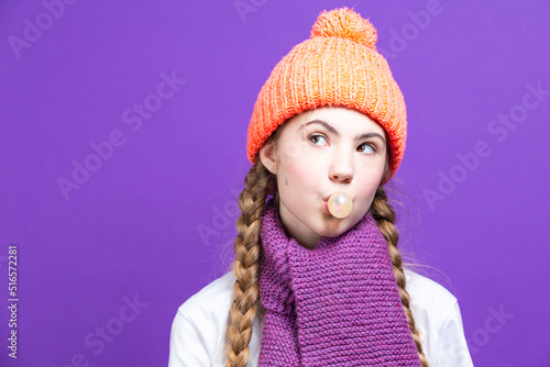 Youth Ideas. One Winsome Girl In Coral Knitted Seasonal Hat Posing With Chewing Gum Bubble In Front of Face Against Purple Seamless Background.