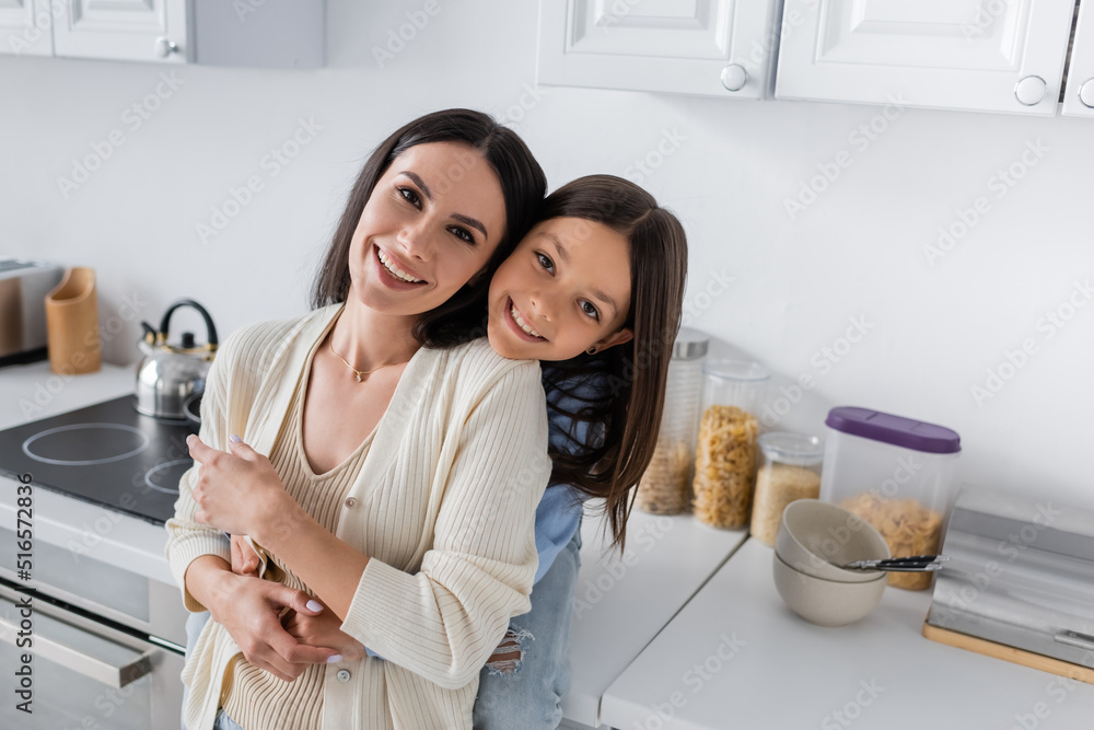 Fototapeta premium happy babysitter with smiling child embracing and looking at camera in kitchen.