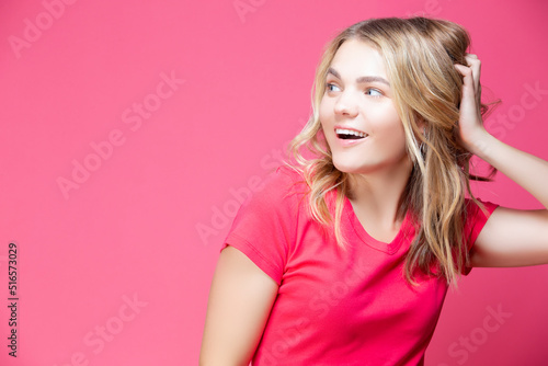 Happy Winsome Caucasian Blond Girl In Coral T-shirt Posing In Summer Shorts While Showing Positive Expression With Hand Lifted Over Coral Pink Background.