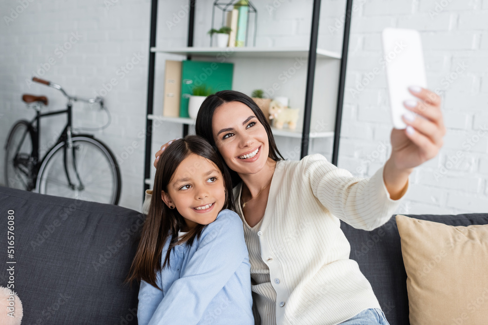 Fototapeta premium smiling babysitter taking selfie on cellphone with girl on sofa at home.