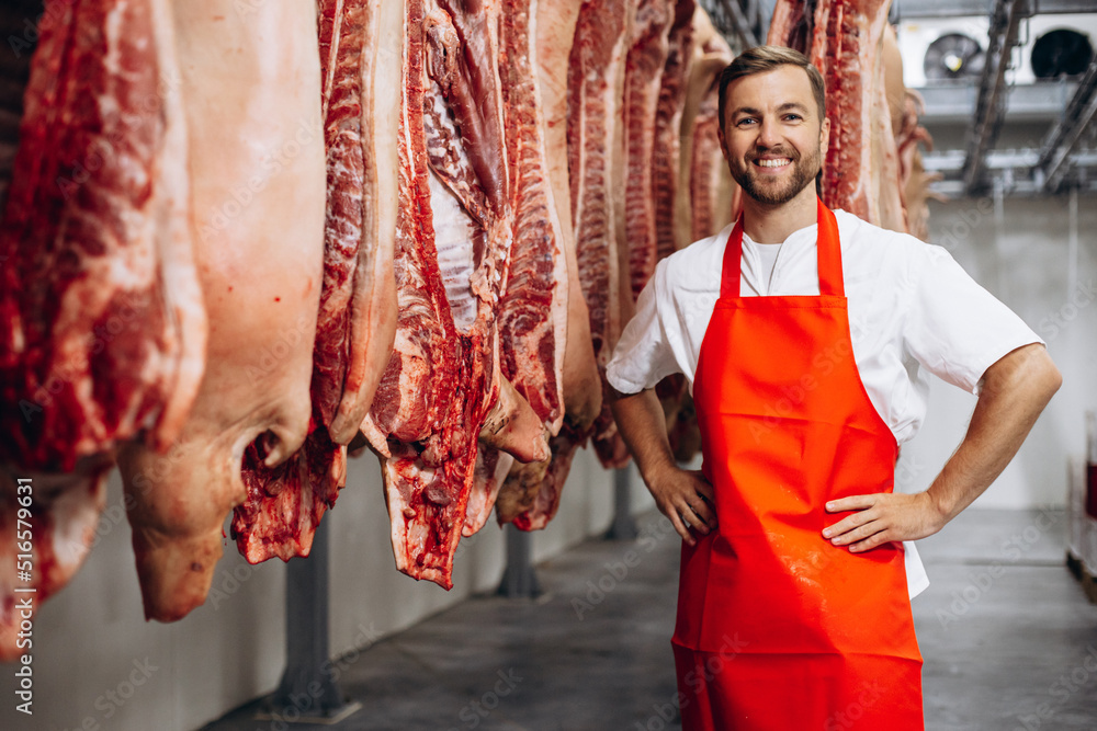 Foto de Young butcher at the meat factory standing by the hanging meat ...