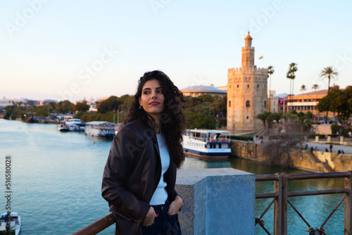 Young and beautiful woman with dark and curly hair is sightseeing in Seville. In the background the river guadalquivir and part of the city in the golden hour. Tourism and holidays concept. photo
