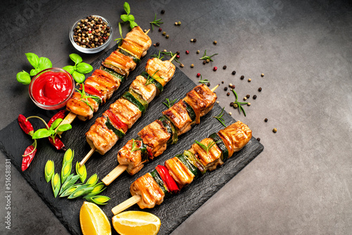 Grilled pieces of chicken meat on skewers.Grilled kebab pieces with vegetables on skewers on a ceramic black board on a dark background with fresh basil leaves.