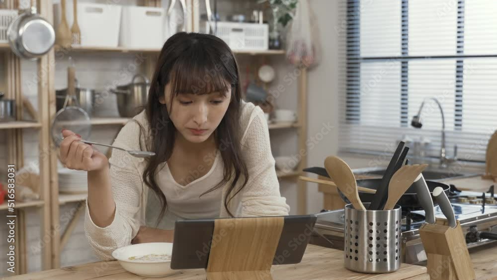 asian woman leaning forward on table is eating oat cereal from the bowl at leisure while reading online on the tablet computer at breakfast in the kitchen. asian woman leaning forward on table is eating oat cereal from the bowl at leisure while reading online on the tablet computer at breakfast in the kitchen.