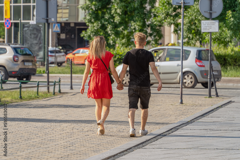 A girl in a red dress and a young man walk together along the city street, holding hands tightly, friendship and love. Young happy couple walking together on a sunny summer evening