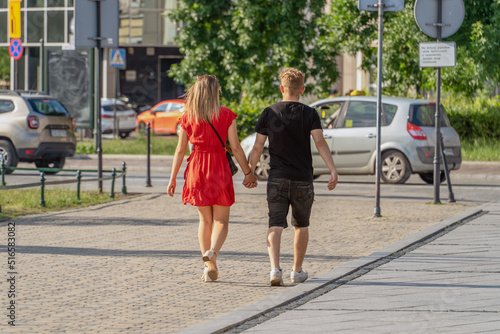 A girl in a red dress and a young man walk together along the city street, holding hands tightly, friendship and love. Young happy couple walking together on a sunny summer evening