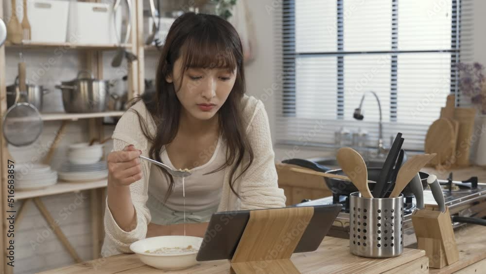 asian girl leaning prone on kitchen table is selecting and watching videos on her touchpad while having a bowl of corn flake with milk for breakfast at home. asian girl leaning prone on kitchen table is selecting and watching videos on her touchpad while having a bowl of corn flake with milk for breakfast at home.