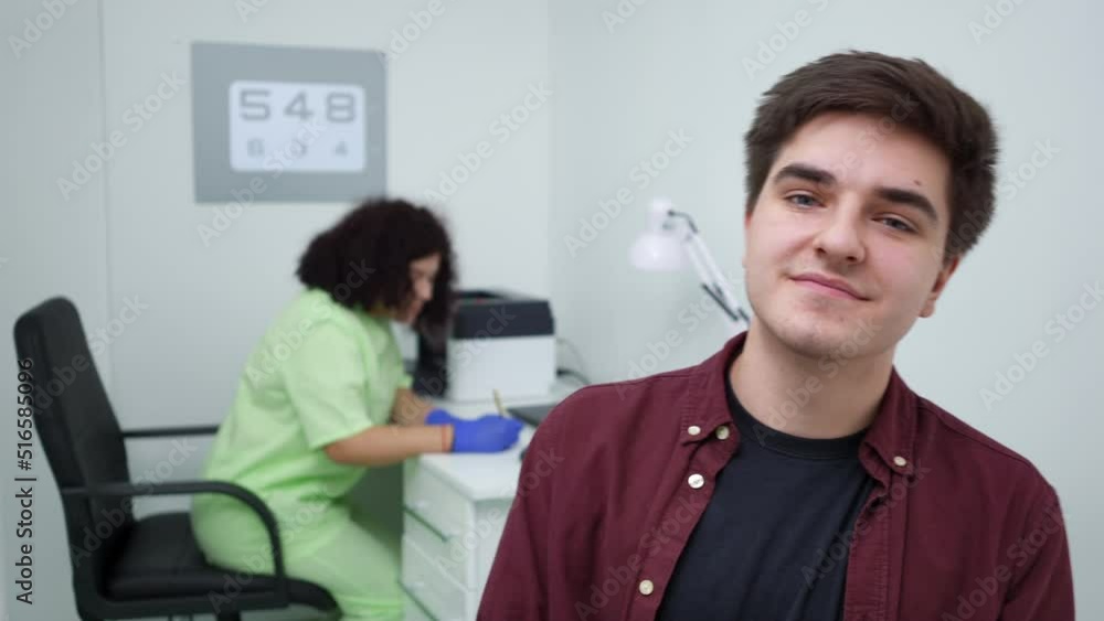 Satisfied patient looking back at blurred doctor turning to camera smiling posing in hospital. Portrait of young Caucasian man happy with appointment standing indoors with nurse filling in paperwork Satisfied patient looking back at blurred doctor turning to camera smiling posing in hospital. Portrait of young Caucasian man happy with appointment standing indoors with nurse filling in paperwork