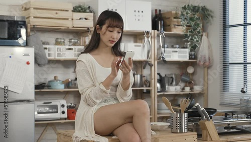 sexy chinese woman sitting on kitchen table with folded leg is checking phone messages in leisure on a tranquil morning at home.