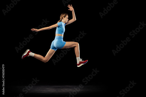 Young sportive girl, long jumper in sports blue uniform performs triple jump isolated on black background. Concept of sport, energy, achievements, motion, speed.