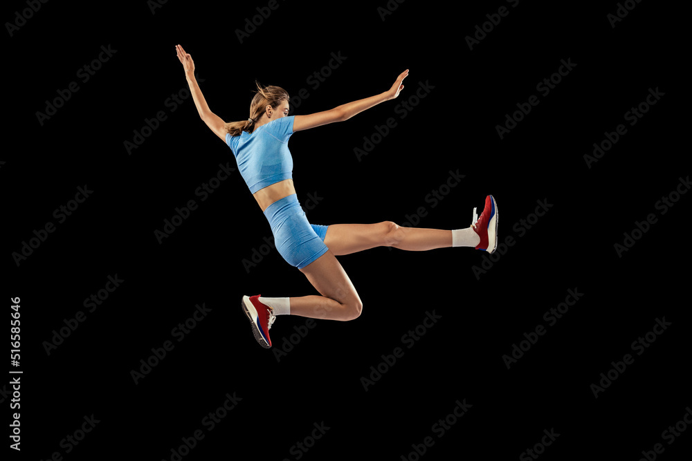 Young sportive girl, long jumper in sports blue uniform performs triple jump isolated on black background. Concept of sport, energy, achievements, motion, speed.