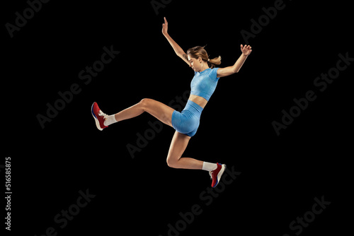Young sportive girl, long jumper in sports blue uniform performs triple jump isolated on black background. Concept of sport, energy, achievements, motion, speed. © master1305