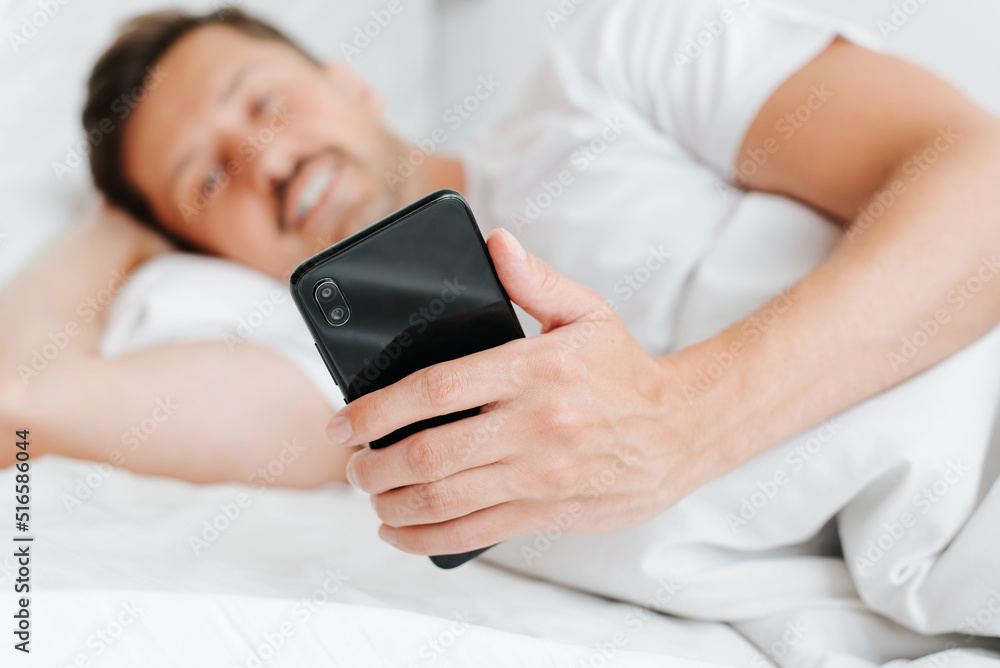 Close-up of happy man holding smartphone while lying in bed in early morning. Pleased guy using mobile phone in bedroom, looking at screen. Selective focus on device Close-up of happy man holding smartphone while lying in bed in early morning. Pleased guy using mobile phone in bedroom, looking at screen. Selective focus on device