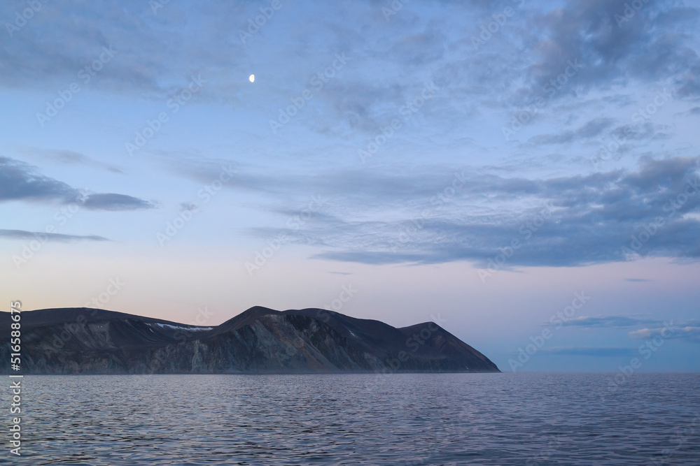 Summer night arctic seascape. View of the mountainous coast. Moon in the sky. Sea voyage to the Arctic. Bering Sea. Eastern coast of Chukotka. Far East of Russia.