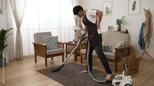 full length of an asian male pulling his back muscles and holding his lower back with a painful groan while vacuuming the living room at home during the day