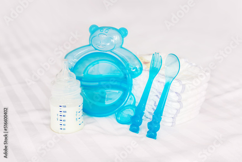 Baby tableware set. milk bottle, feeding nibbler, blue cutlery, spoon, fork, diapers, disposable diapers on the white bed background. front view