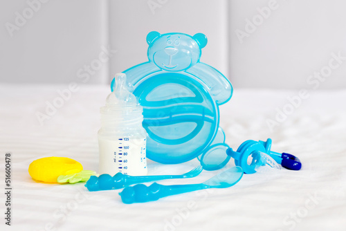 Baby tableware set. milk bottle, feeding nibbler, blue cutlery, spoon, fork, diapers, toy, disposable diapers on the white bed background