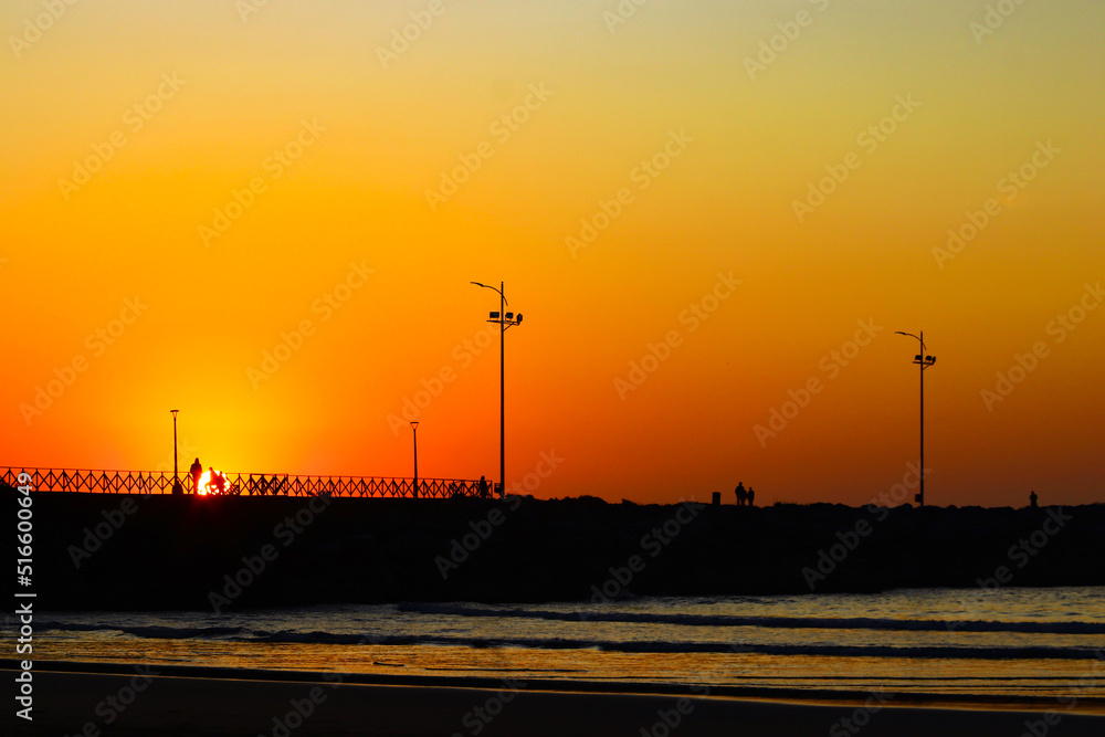Nascer do sol em Itajaí, Brasil, com céu laranja e amarelo refletindo no mar com o farol ao fundo e silhueta de pessoas. 