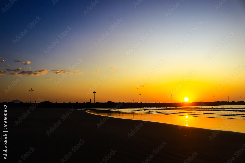
Sunrise in Itajaí, Brazil, with orange and blue sky reflecting in the sea and lighthouse in the background in a silhouette