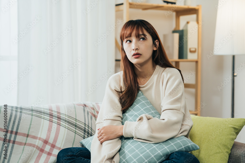 unhappy chinese girl sitting with a pillow in arms on the living room sofa is looking away with boredom while staying at home alone during quarantine. unhappy chinese girl sitting with a pillow in arms on the living room sofa is looking away with boredom while staying at home alone during quarantine.
