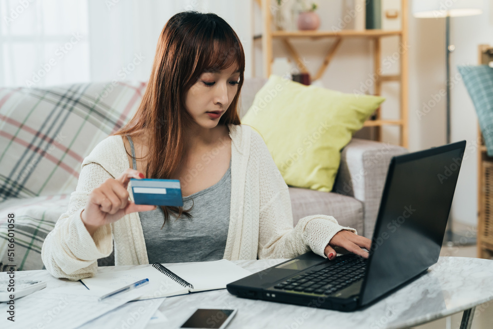 portrait attractive asian woman is entering numbers on the computer while paying tax online with a credit card in the living room at home. portrait attractive asian woman is entering numbers on the computer while paying tax online with a credit card in the living room at home.