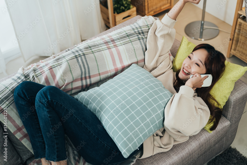 Obraz premium view from top happy asian lady talking on the phone is cheering for good news with a lifting arm while lying comfortably on the living room couch at home.