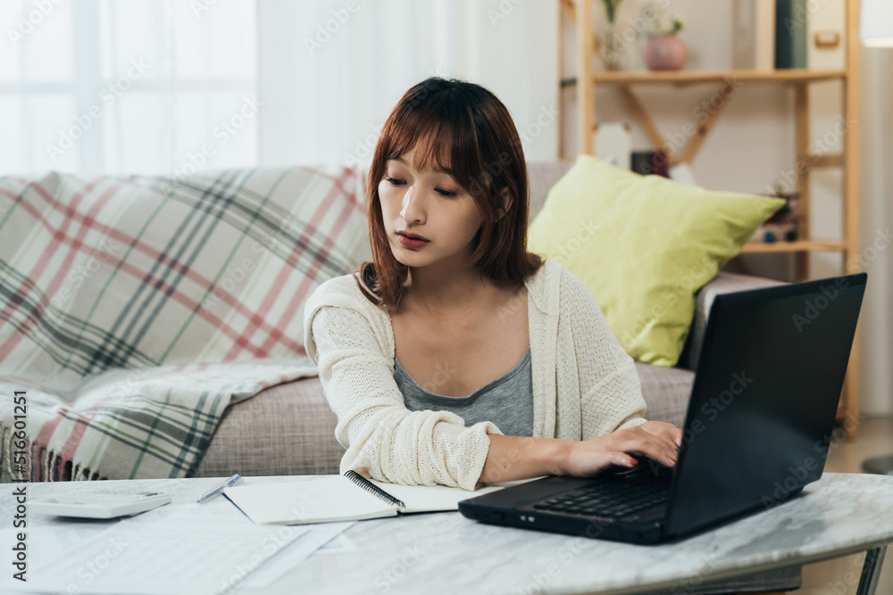 portrait beautiful asian female is looking at the financial document and entering information on the laptop while filing tax online in the living room at home. portrait beautiful asian female is looking at the financial document and entering information on the laptop while filing tax online in the living room at home.