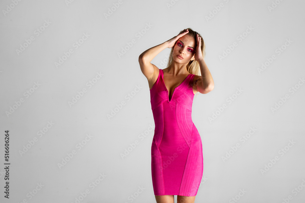 Fototapeta premium Front image of a sexy blonde female model in pink slim dress isolated on grey background. Space for text.