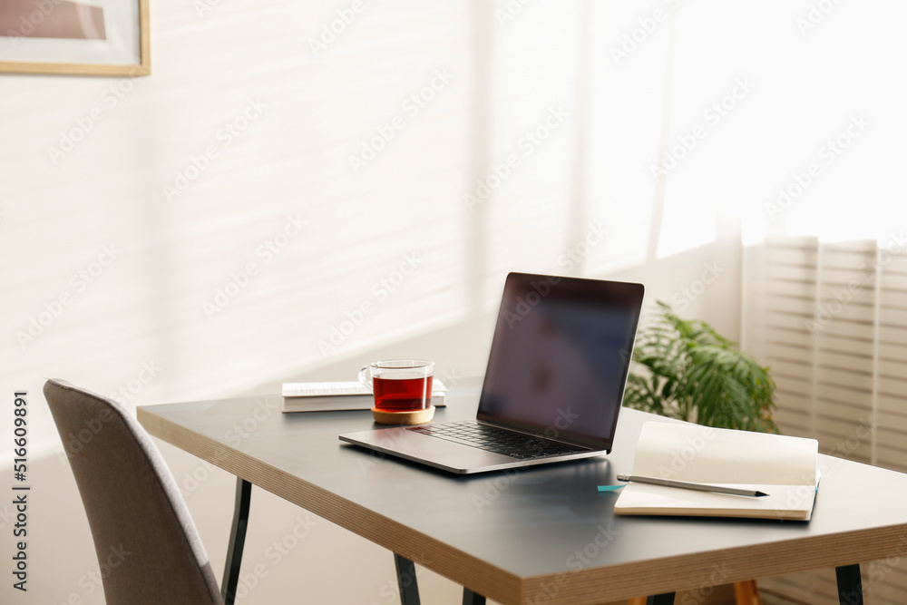 Home office concept. Designated work from home area near the window. Modern laptop with blurred screen and glass cup of tea on table. Close up, copy space, interior background. Home office concept. Designated work from home area near the window. Modern laptop with blurred screen and glass cup of tea on table. Close up, copy space, interior background.