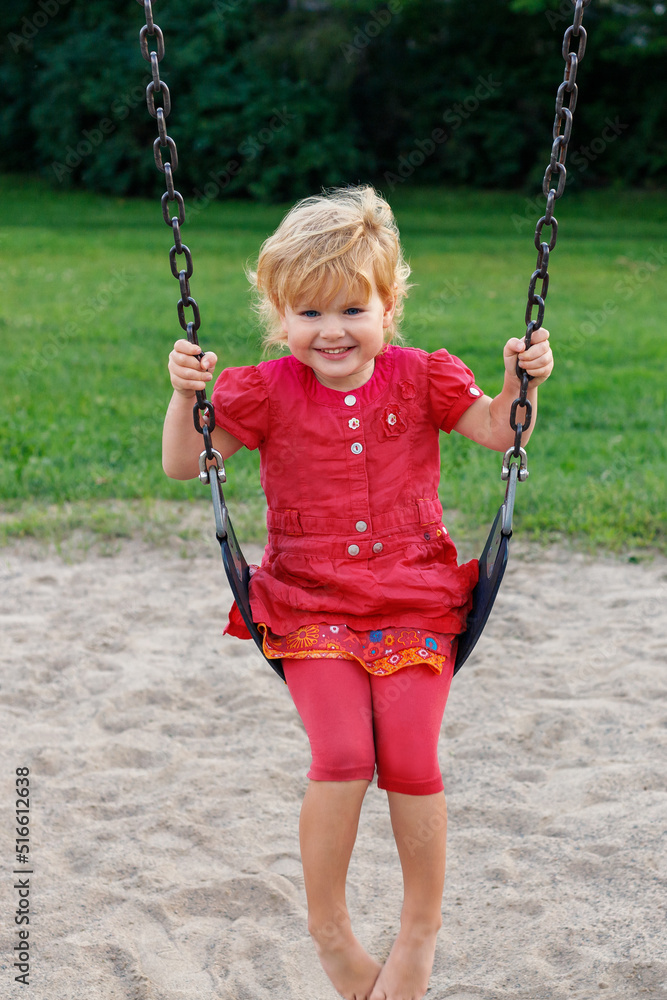 Smiling child sitting on swing. Happy kid playing in park. Girl swinging at playground in summer.