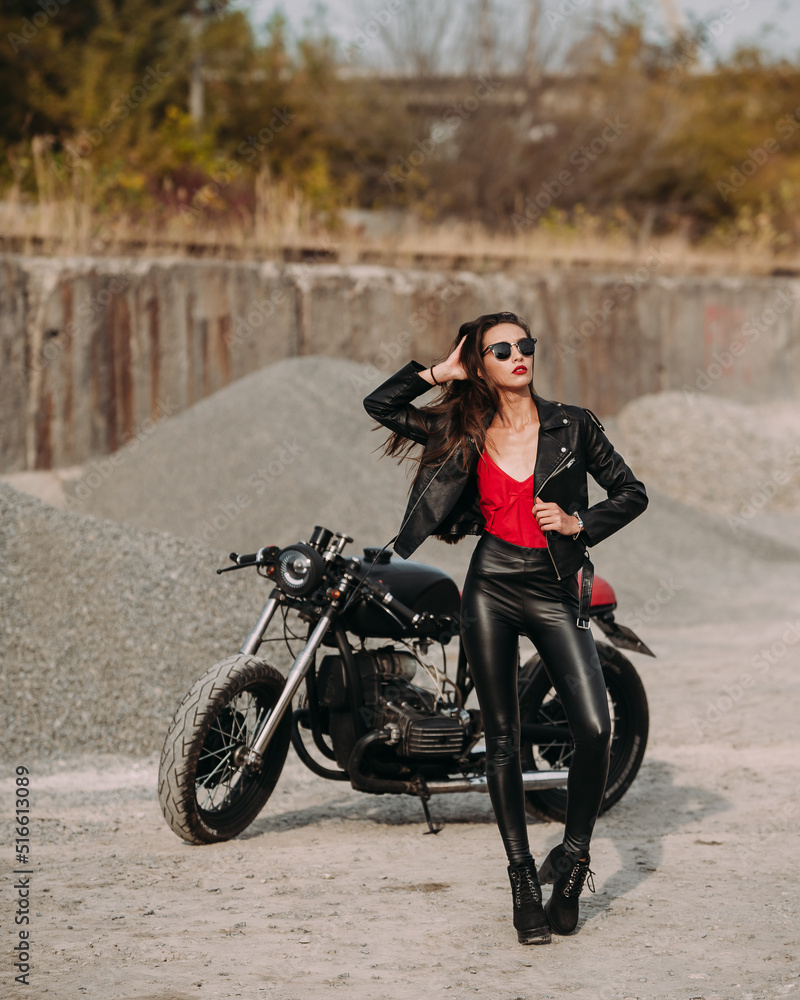 beautiful girl with long dark hair red lips in black jacket and black pants standing near black vintage motorcycle in industrial zone beautiful girl with long dark hair red lips in black jacket and black pants standing near black vintage motorcycle in industrial zone