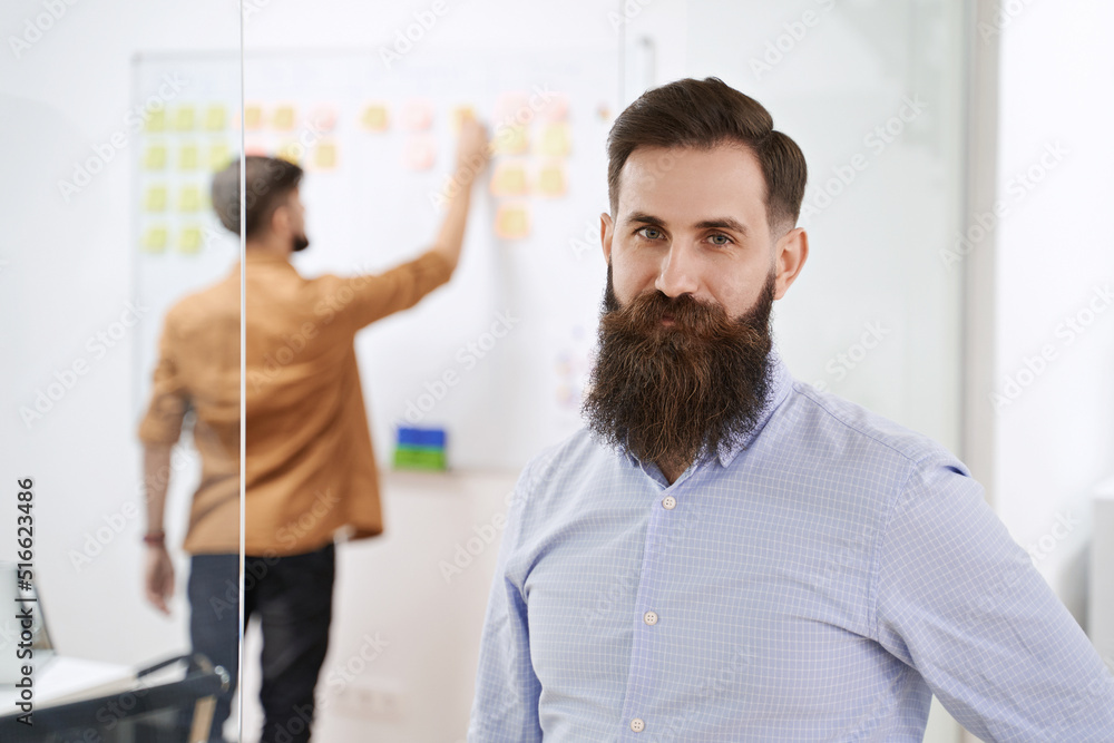 Happy smiling bearded senior developer or manager in modern IT office. Another professional working with scrum desk on background. Successful technology project or startup concept. High quality image.