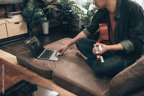 young man relax and playing guitar while sitting on sofa bed in living room at home. Music create melody song, lyrics on laptop and practice concept.