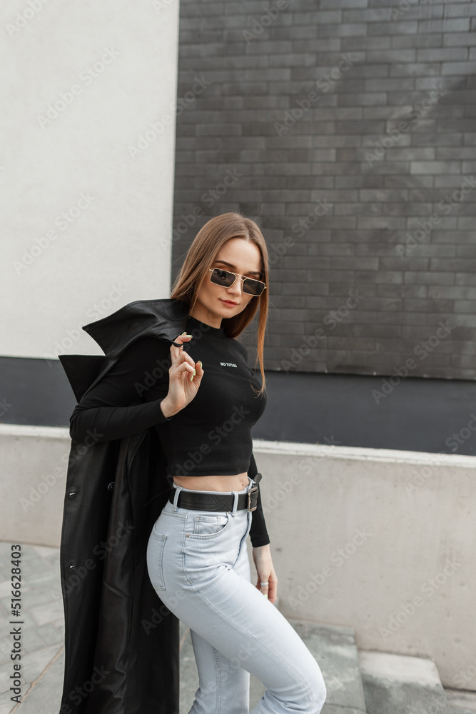 Naklejka premium Fashionable beautiful young woman model with sunglasses in fashionable black clothes with black T-shirt and vintage high waist jeans with coat walking outside