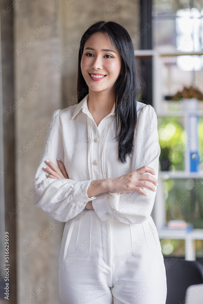 Portrait of a smiling young Asian businesswoman leaning against a table while standing working alone in a large modern office Portrait of a smiling young Asian businesswoman leaning against a table while standing working alone in a large modern office