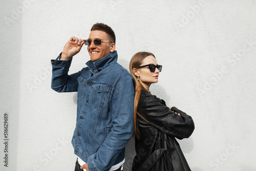 Fashionable young couple in trendy urban casual clothes posing near a wall on the street. Fashion handsome happy hipster man and his girlfriend wearing vintage sunglasses in stylish denim outfit