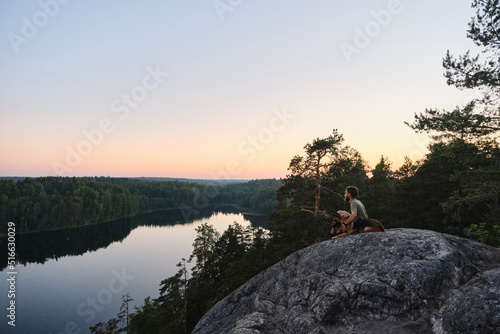 Concept of traveling in Russia. Young man is sitting on rock with dog and admiring nature and river below. German shepherd dog travels with owner in mountains. Republic of Karelia.
