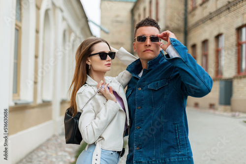 Stylish cool beautiful couple of models with trendy sunglasses in fashionable casual denim clothes and a fashion bag are walking on the street near a vintage building