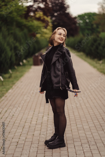 Cute happy girl with glasses in a black jacket and dress walks in the park alone in autumn. Pretty young girl with blond hair in black walks alone in the park and poses on the empty road. 