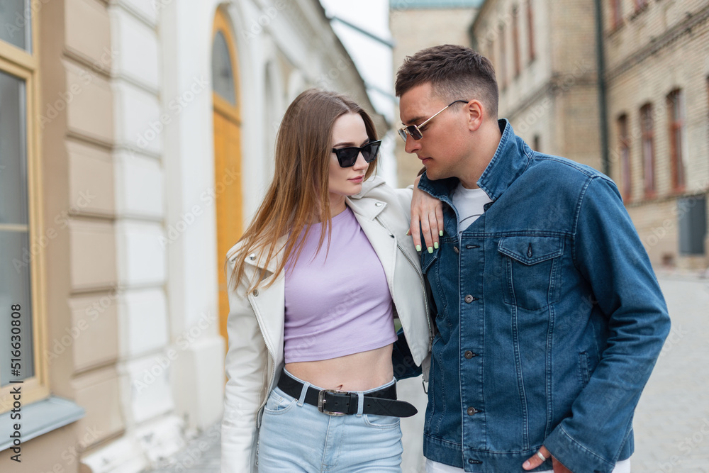 Fashionable young beautiful hipster couple with sunglasses in stylish spring urban denim clothes walks in the city