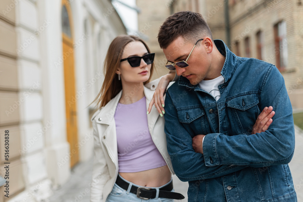 Cool stylish handsome man model with hairstyle and sunglasses in a trendy jeans jacket and a young trendy hipster girl in fashion clothes look are walking in the city