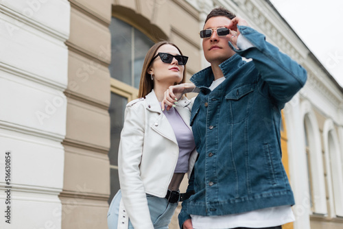 Cool fashionable young beautiful couple girl and boy with sunglasses in fashion casual outfit with denim jacket and leather jacket walks in the city. Trendy stylish clothes looks