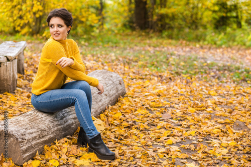 Beautiful young woman in a yellow knitted sweater and blue jeans sits on a log in an autumn park