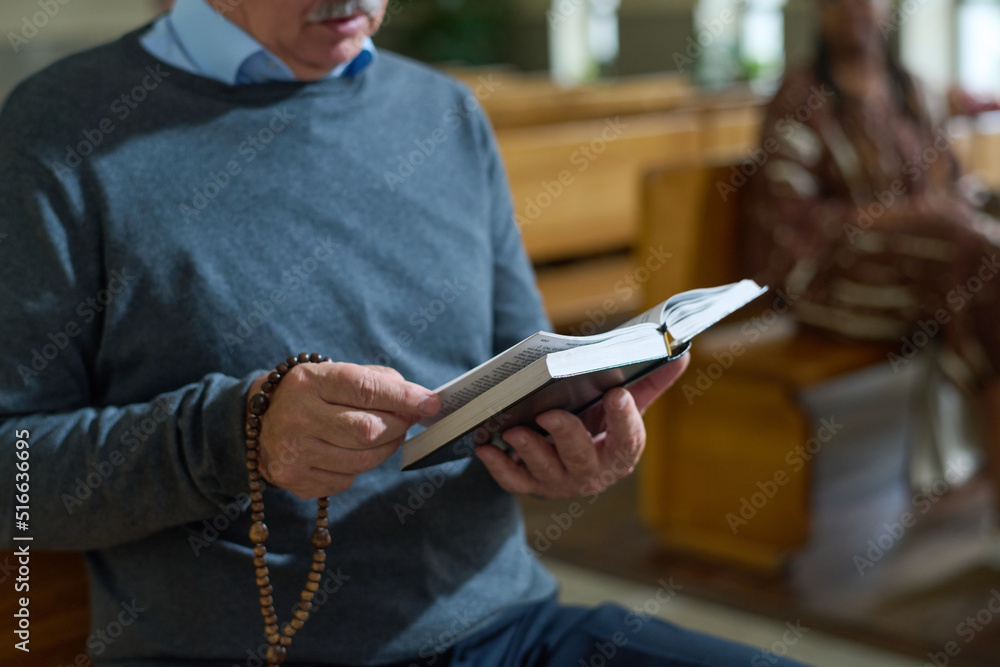 Hands of aged man holding open Bible and wooden rosary beads while reading verses from Gospel or some other book during sermon