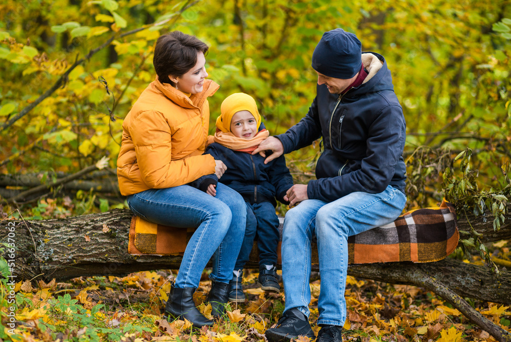 Mother, father and little son sit on a plaid on a log in the autumn park and spend time together Mother, father and little son sit on a plaid on a log in the autumn park and spend time together