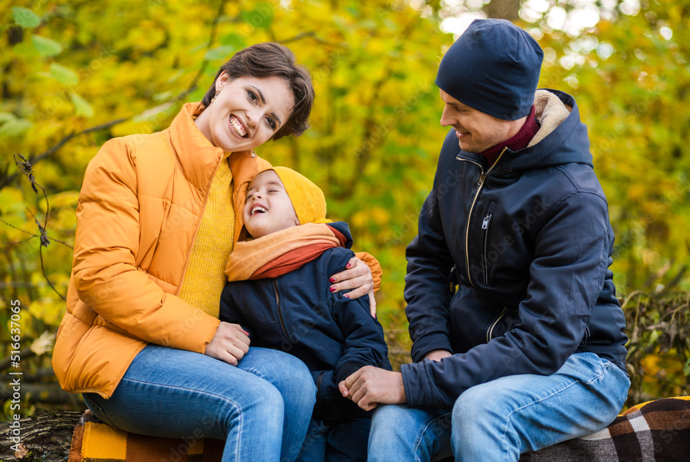 Mother, father and little son sit on a plaid on a log in the autumn park and spend time together