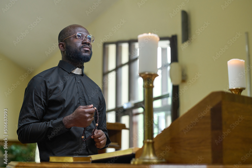 African American priest in black casualwear holding rosary beads during pray while standing by pulpit with two burning candles - obrazy, fototapety, plakaty 